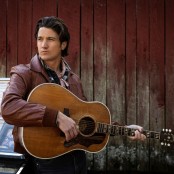 A man wearing a brown leather jacket holding an acoustic guitar
